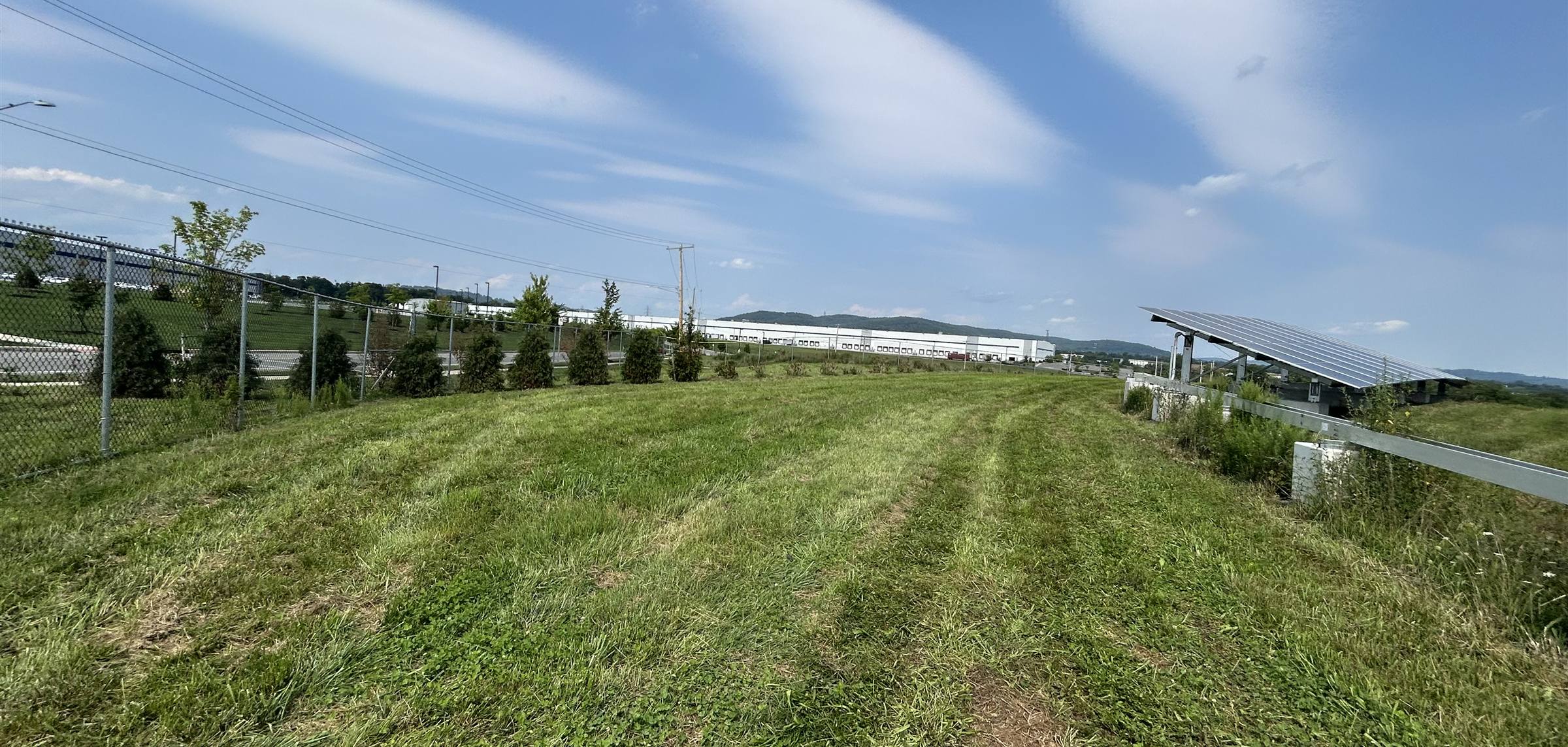 Freshly mowed ground running along a fence line beside a ground-mount solar array under a wide blue sky
